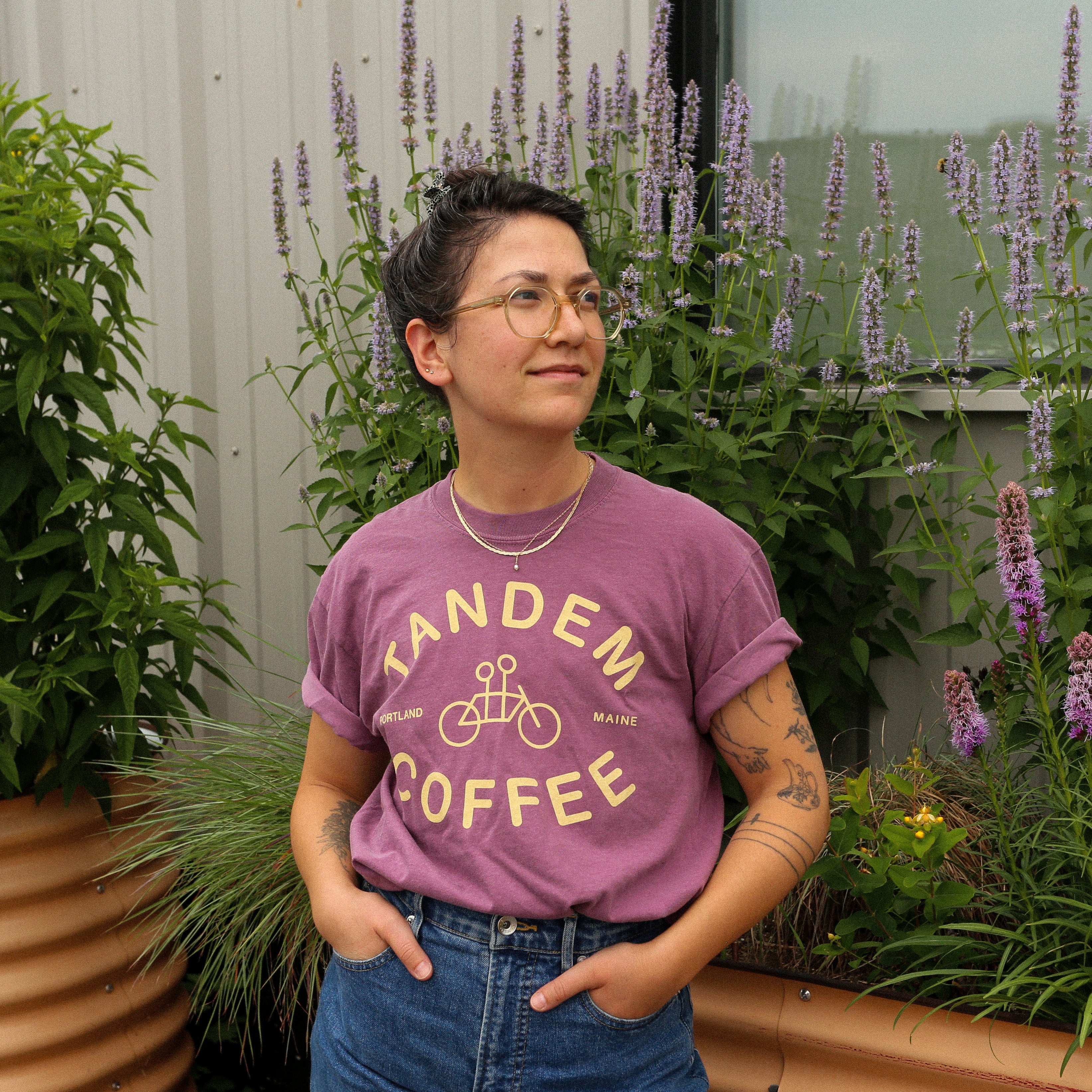 A person with short dark hair, glasses, and tattoos stands in front of tall flowers and a metal building, wearing the Berry Logo Tee by Tandem Coffee Roasters with jeans, hands in their pockets, looking off to the side.