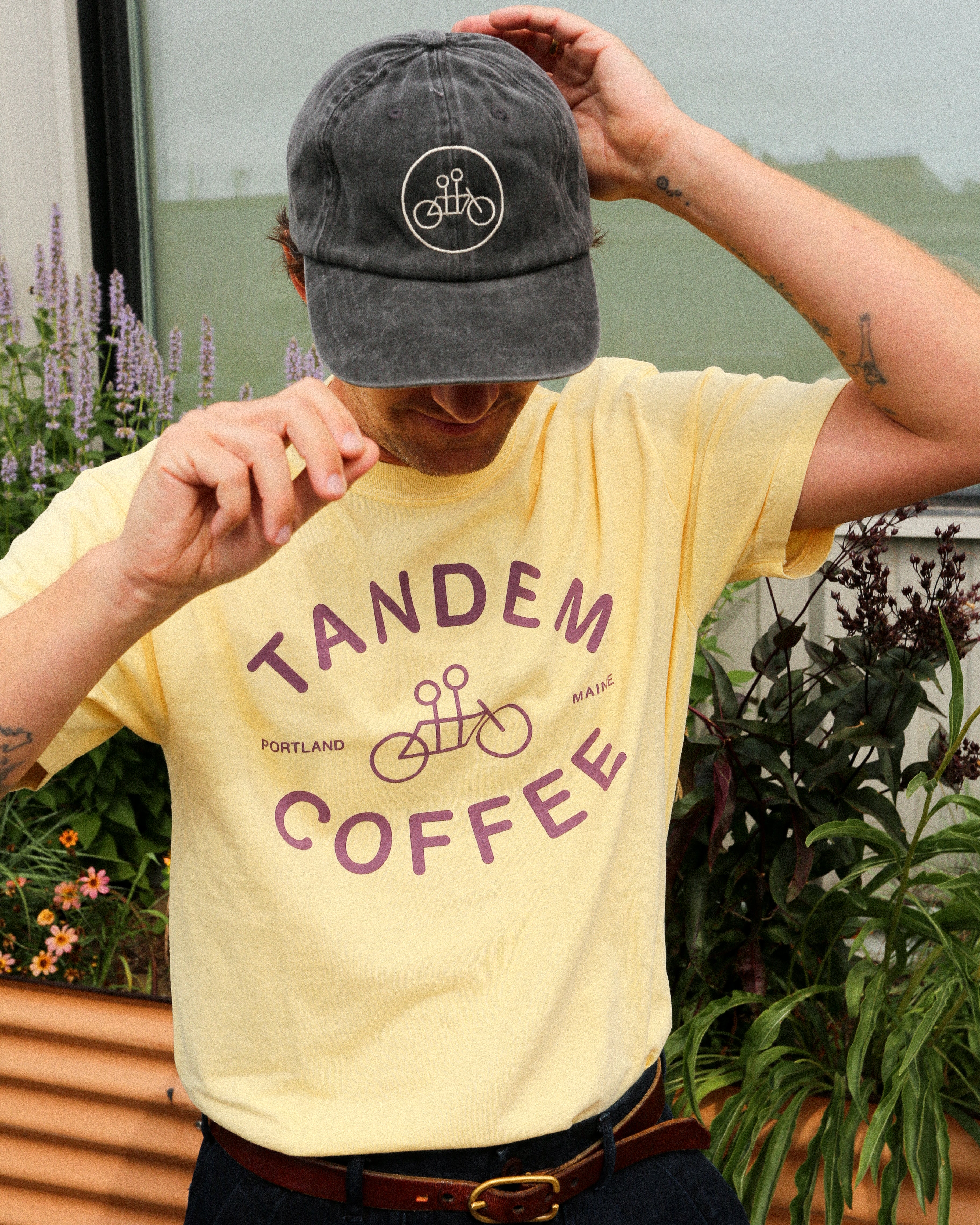 A person wearing a Butter Yellow Logo Tee from Tandem Coffee Roasters and a dark cap stands outdoors among greenery, adjusting their hat with one hand while looking down.
