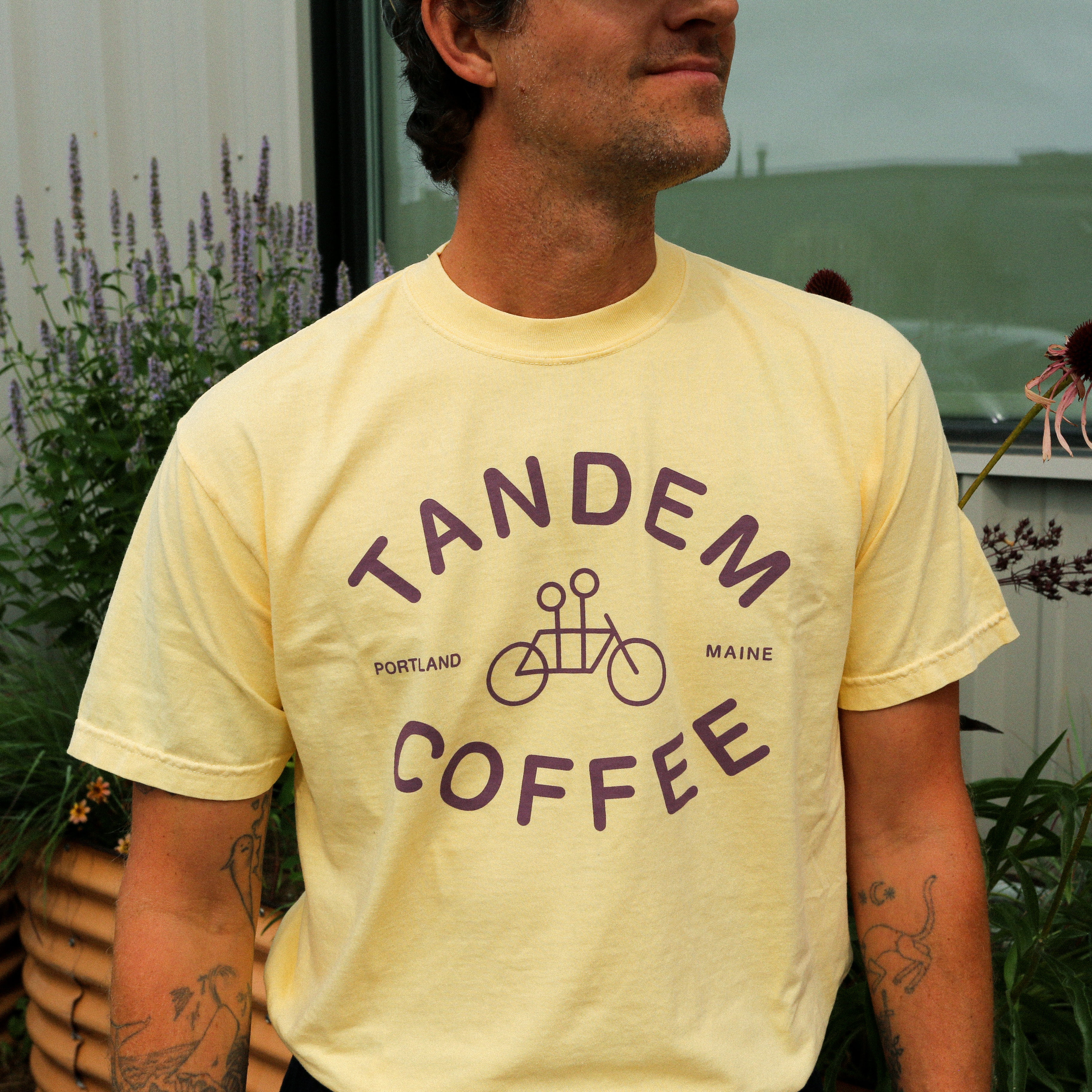 A person wearing a Butter Yellow Logo Tee from Tandem Coffee Roasters, featuring a tandem bicycle graphic, stands outside near green plants and purple flowers.