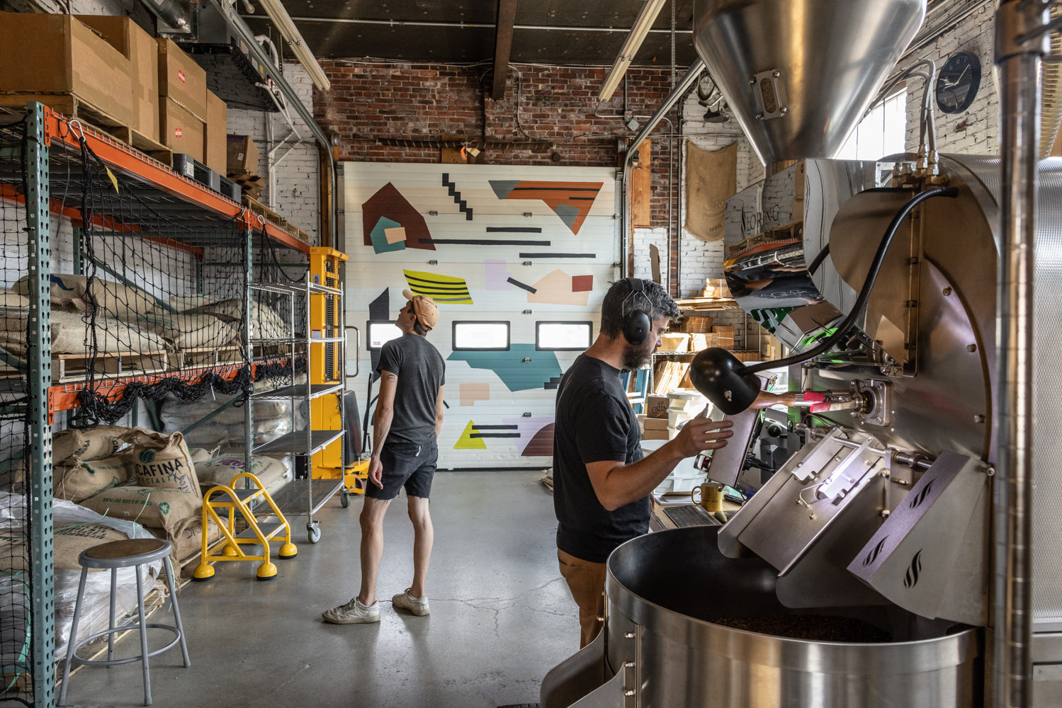 Two people working in a coffee roastery. One person, wearing a cap, stands near tall shelves filled with large sacks and equipment. The other person, wearing headphones, operates a coffee roasting machine. A colorful, geometric mural decorates the back wall of Tandem Coffee in Portland, Maine.