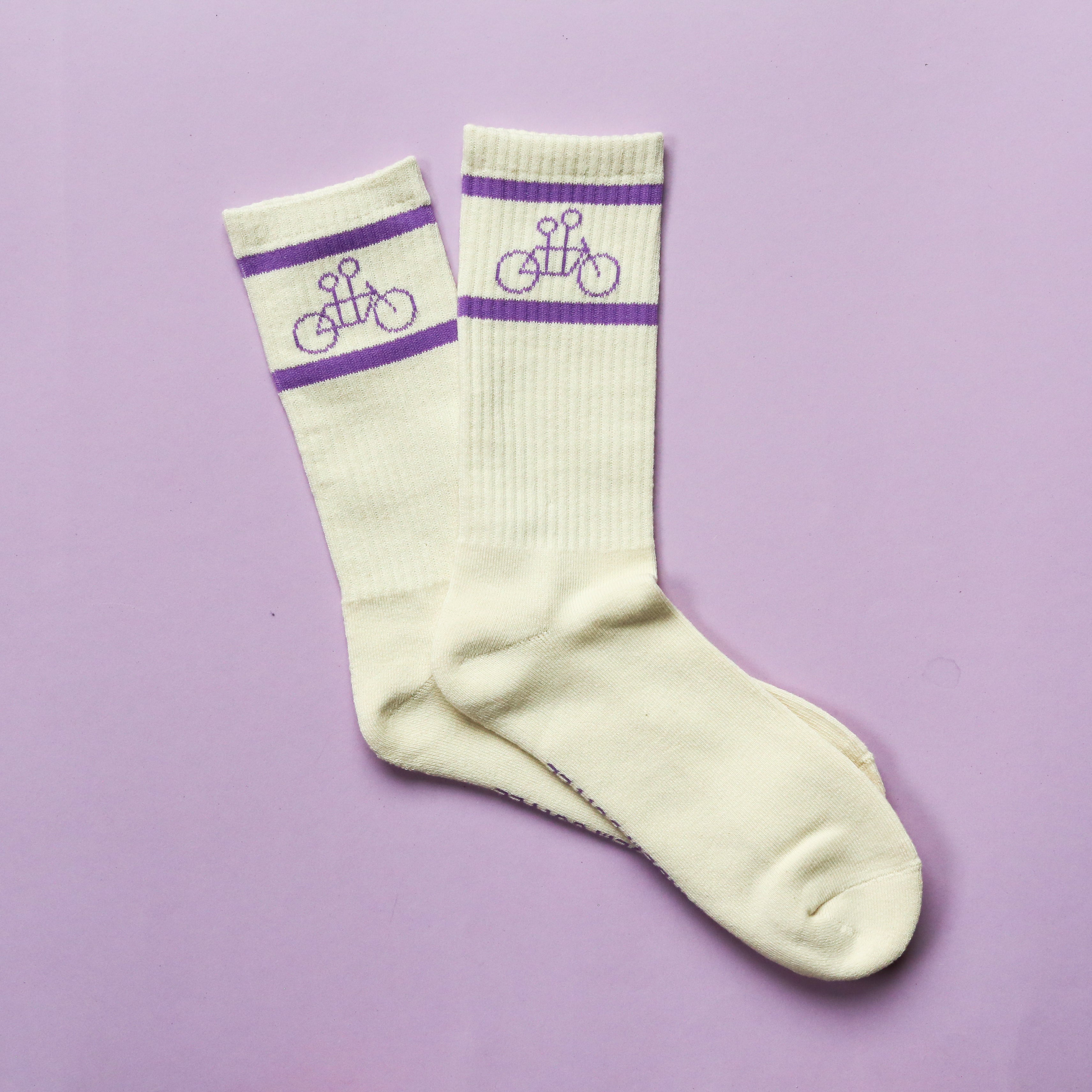 The Tandem Socks by Tandem Coffee Roasters are white cotton socks with purple stripes and bicycle designs near the top, shown on a light purple background. One size fits most—perfect for Tandem Coffee fans.