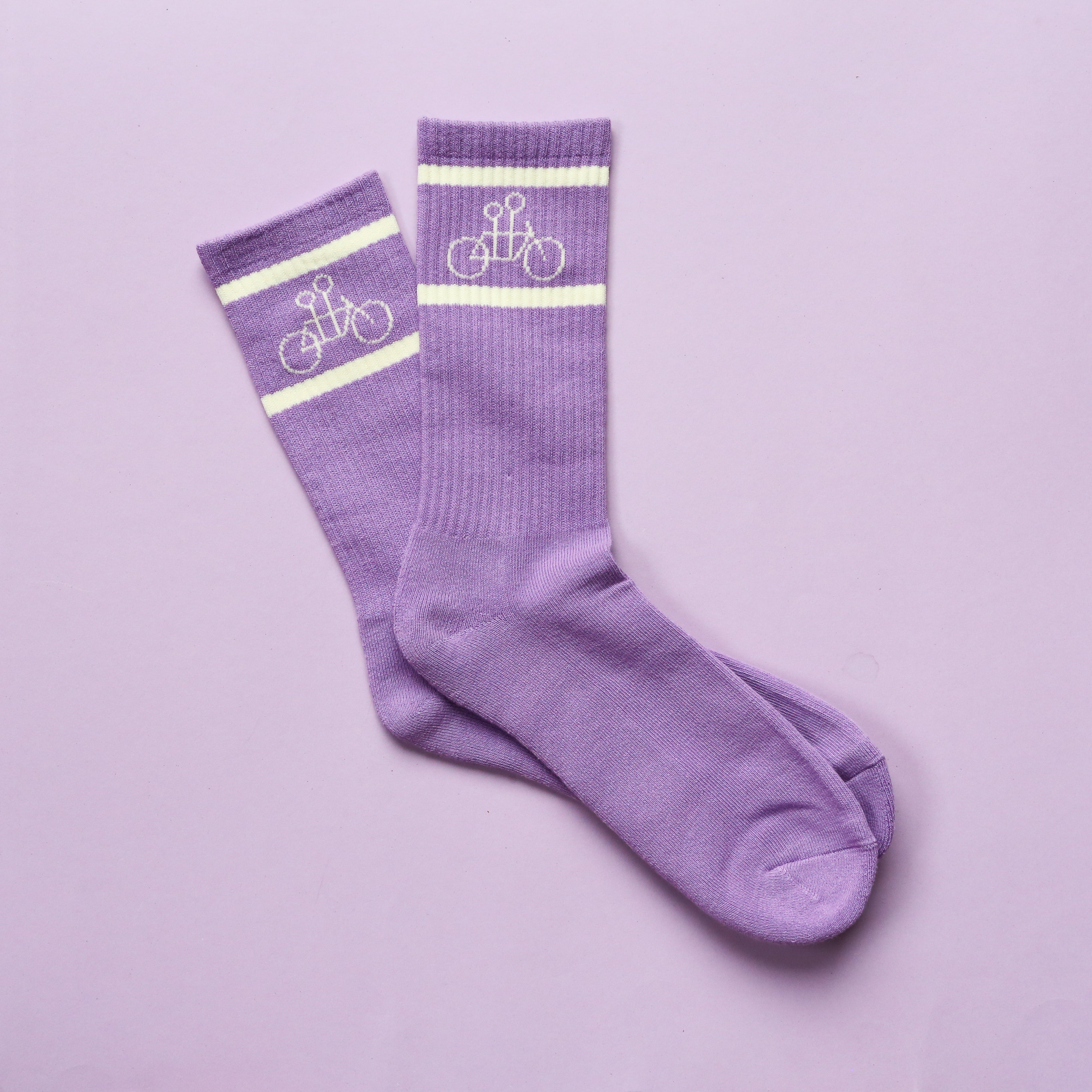 Two purple The Tandem Socks by Tandem Coffee Roasters with white stripes near the top and a white bicycle design above, placed side by side on a matching purple background.