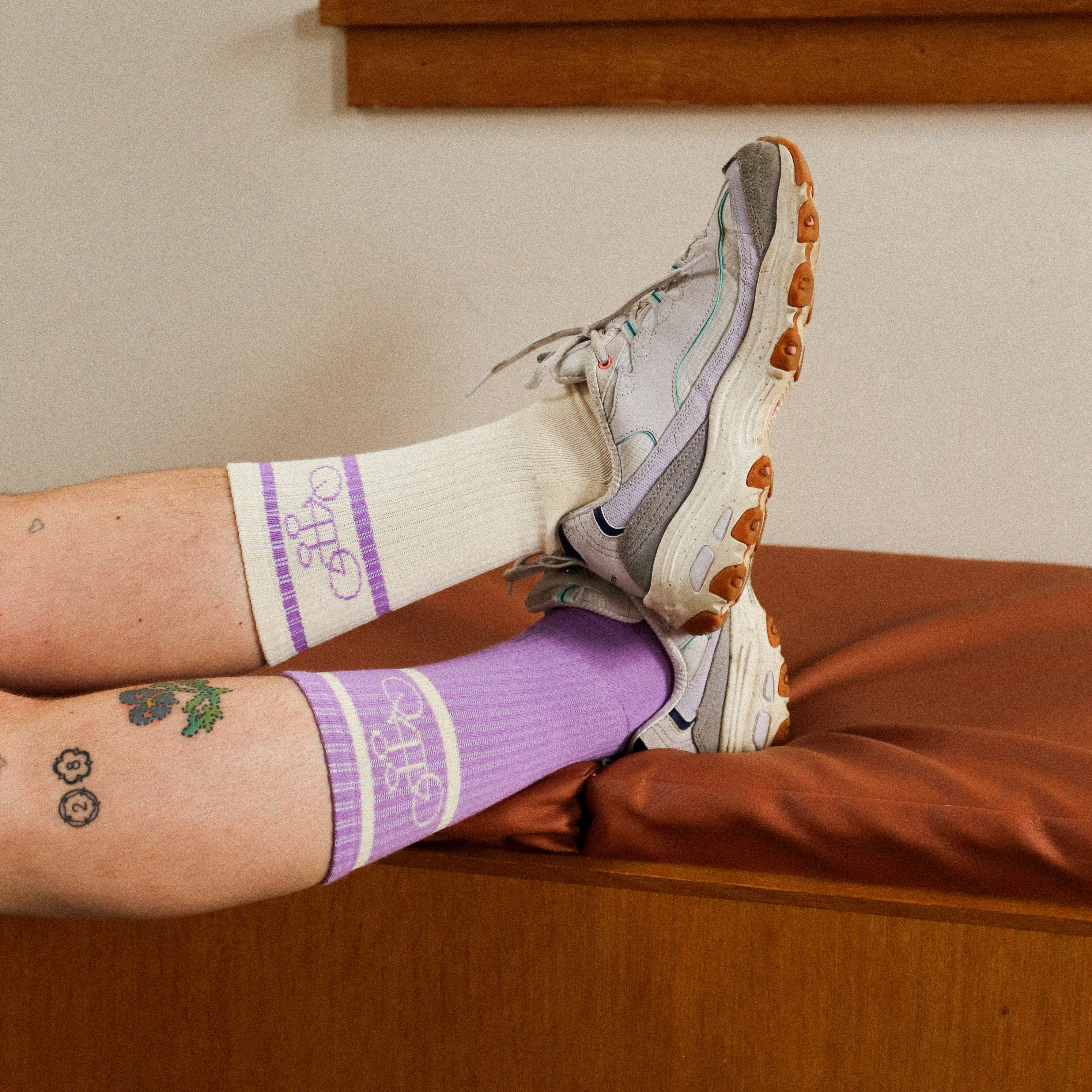 Tattooed legs crossed at the ankles, wearing The Tandem Socks by Tandem Coffee Roasters—white and purple cotton socks with a logo—paired with gray sneakers and resting on a brown bench near a wooden wall.