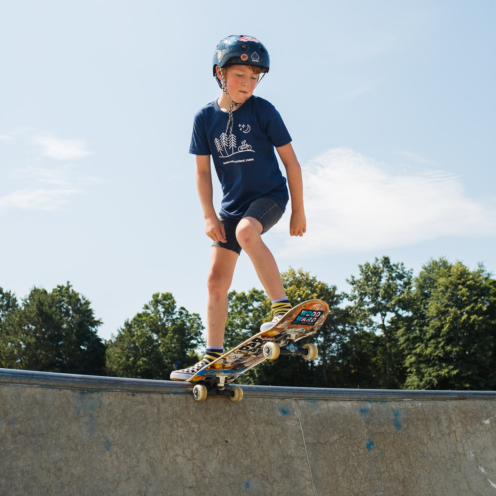 A young boy wearing a helmet and a Tandem Kids Tandem Moon Tee is skateboarding at the edge of a concrete skate park ramp on a sunny day.