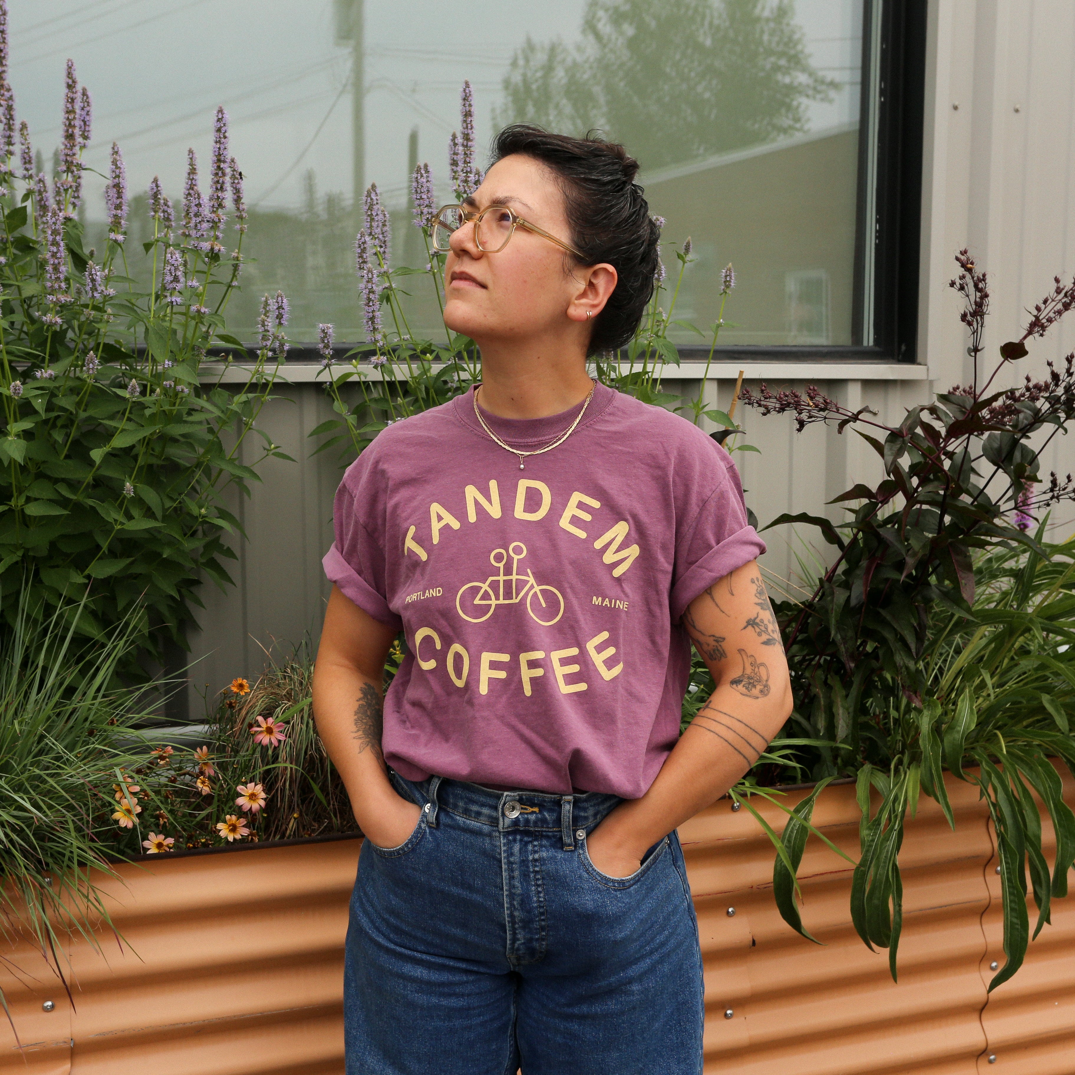 A person with short hair and glasses stands in front of tall plants, wearing the Tandem Coffee Roasters Berry Logo Tee made from 100% cotton, paired with blue jeans. Their hands are in their pockets as they look upward thoughtfully.