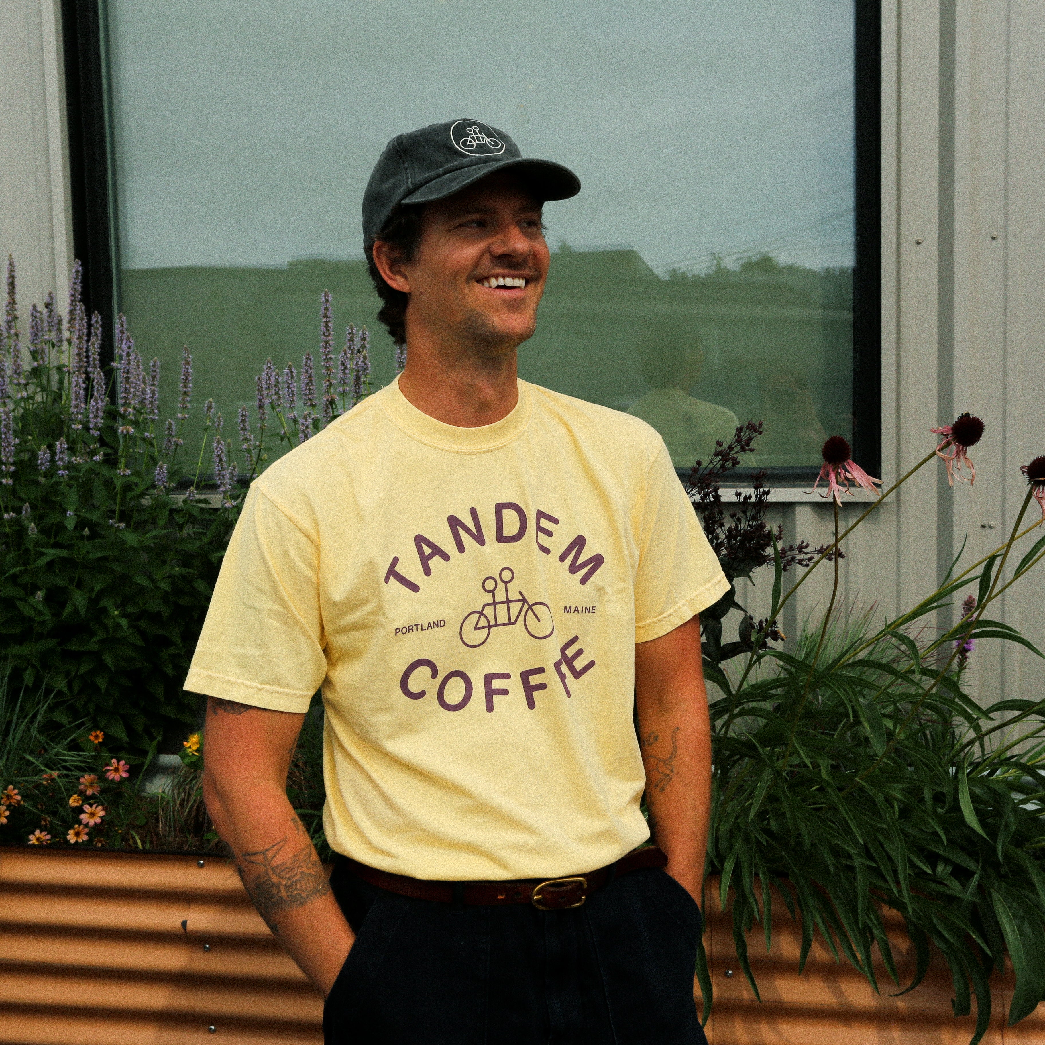 A smiling person wearing a Butter Yellow Logo Tee from Tandem Coffee Roasters and a dark cap stands in front of a raised garden bed filled with flowers and greenery, with a building and window in the background.