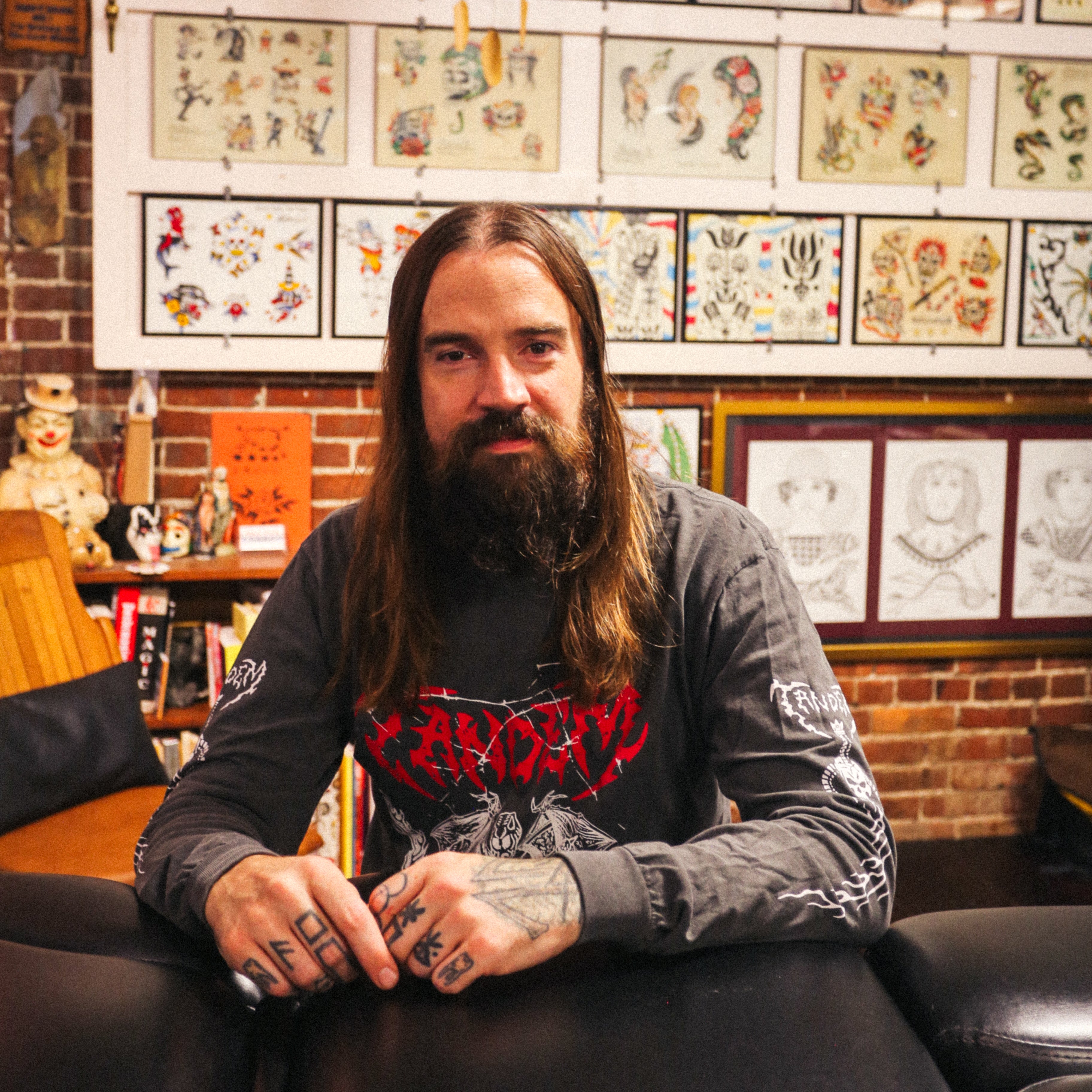 A bearded man with long hair sits at a table in Gold Star Tattoo, surrounded by tattoo flash art on brick walls. He has tattoos on his fingers and wears a dark Tandem Coffee Roasters Tandem Metal Long Sleeve tee.