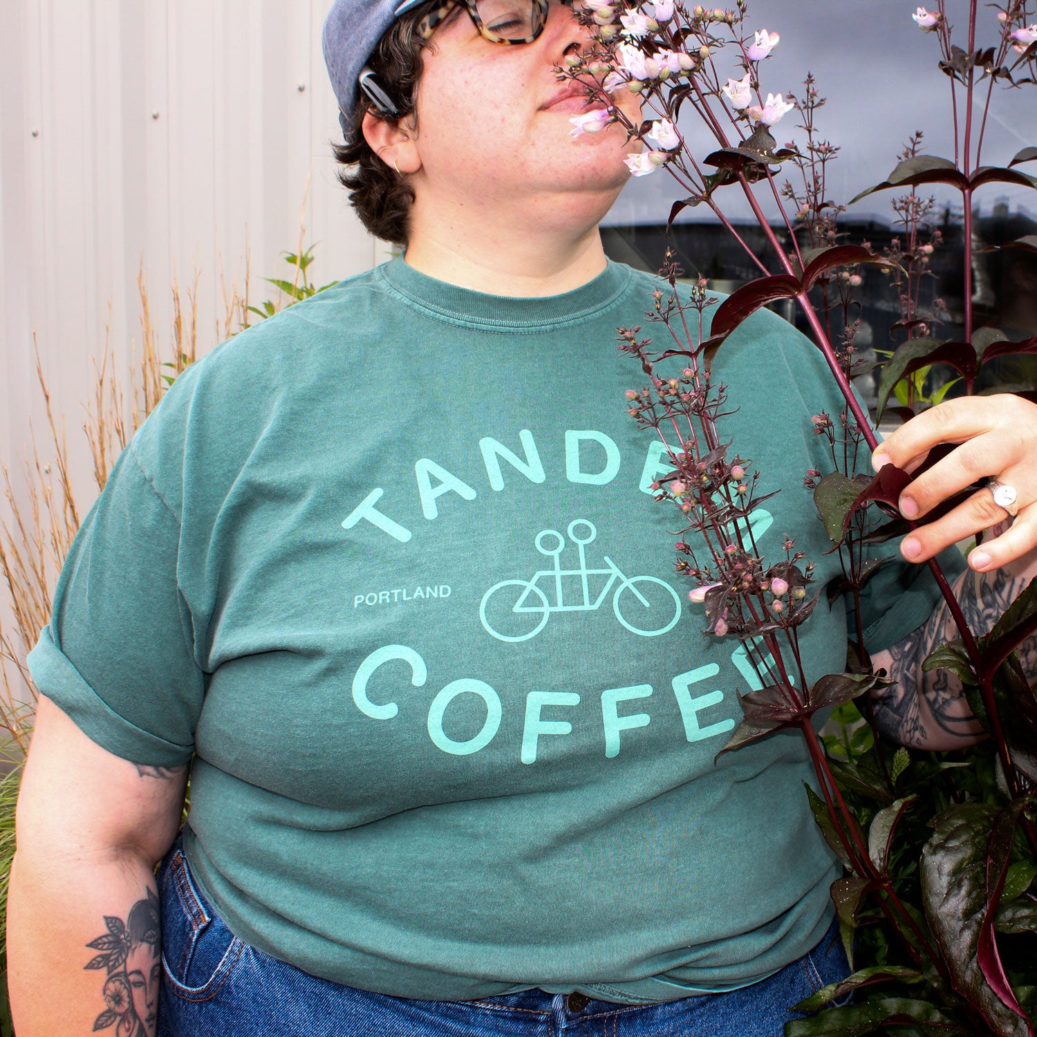 A person in glasses and a cap stands outside, wearing a blue Tandem Coffee Roasters Blue Spruce Logo Tee, holding and smelling a tall plant with purple leaves and small pink flowers.