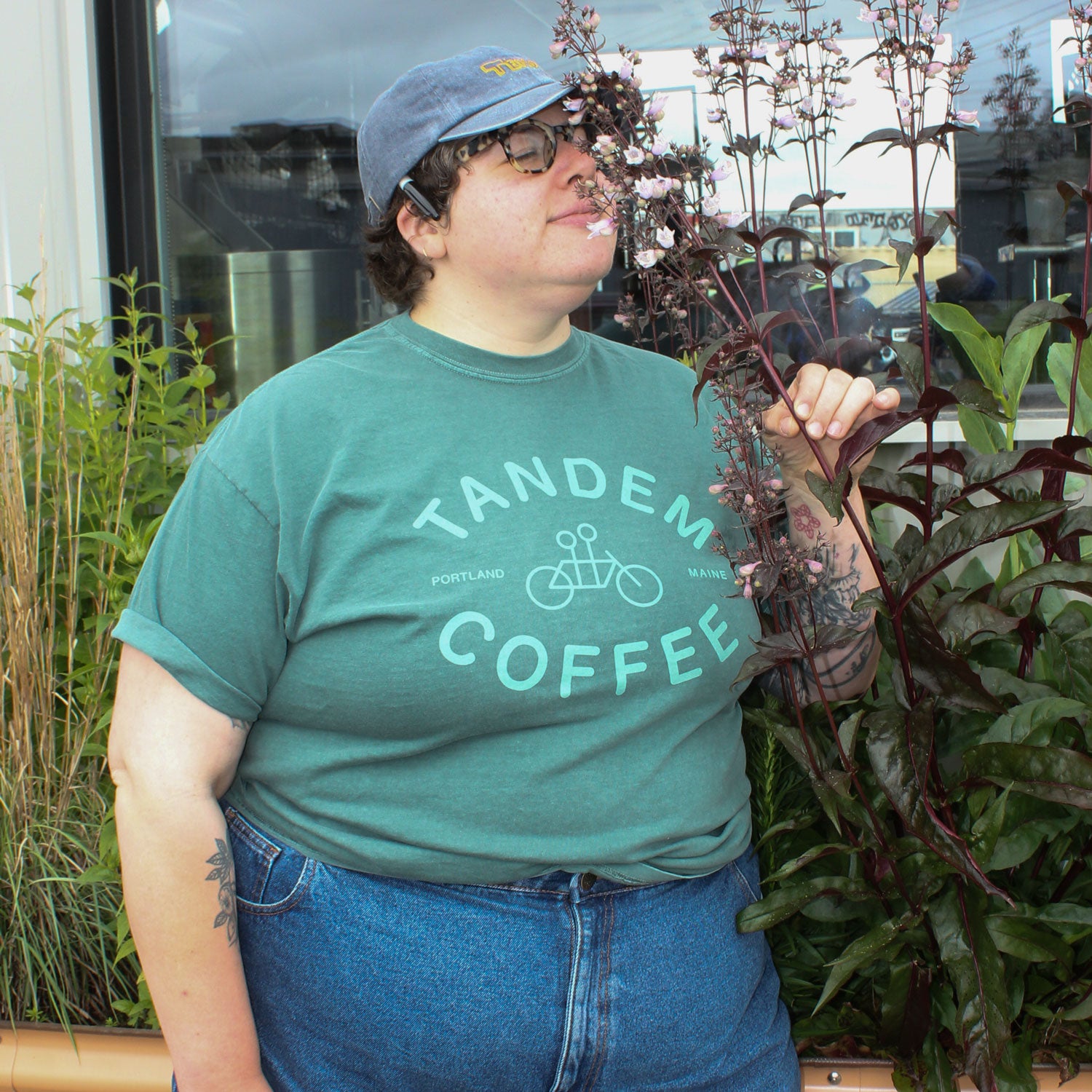 A person in a green Tandem Coffee Roasters Blue Spruce Logo Tee, blue jeans, and a blue cap smells a tall flowering plant outside near a building with glass windows.
