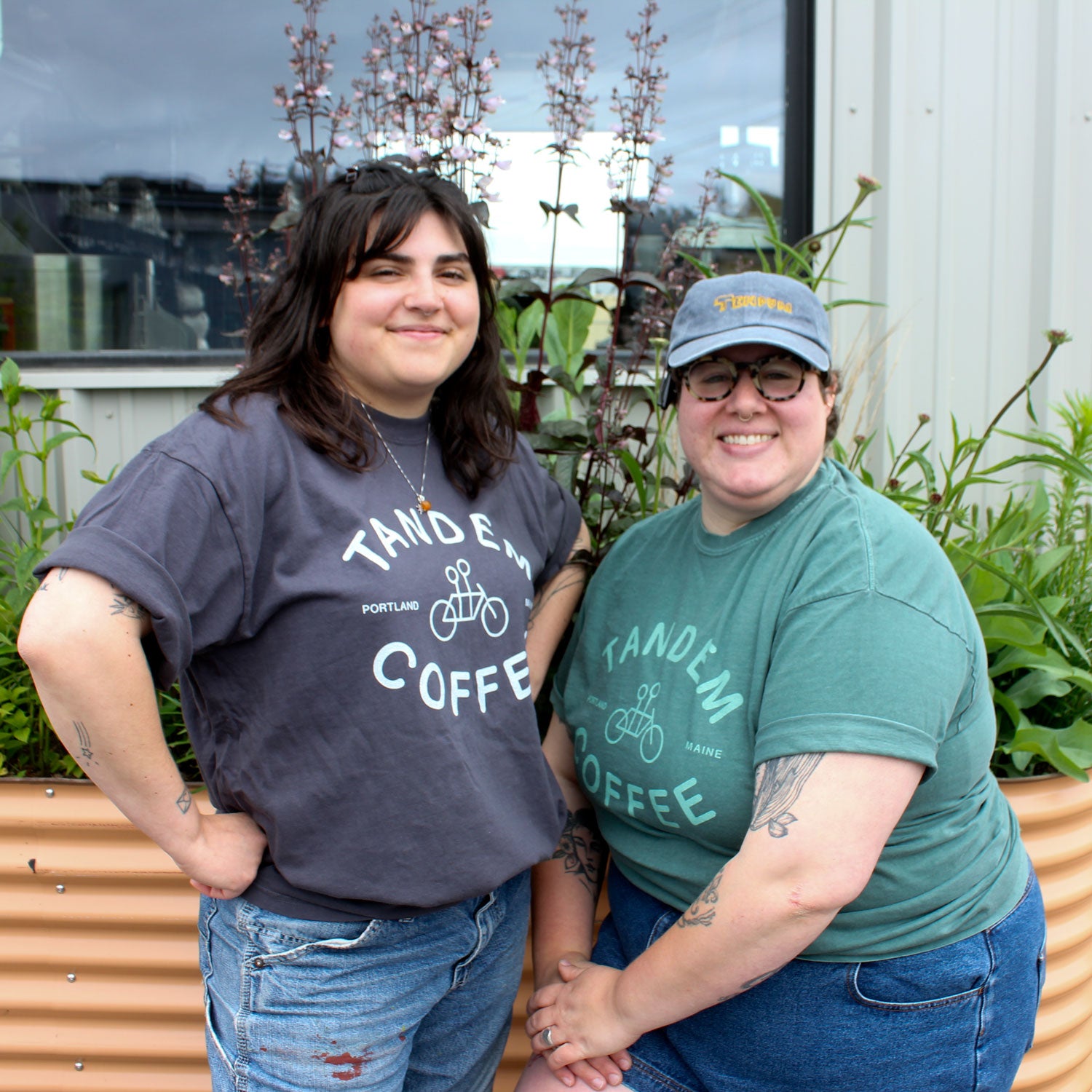 Two people in Tandem Coffee Roasters Blue Spruce Logo Tees stand smiling outside near a raised garden bed, with lush plants and a blue spruce visible by the building in the background.