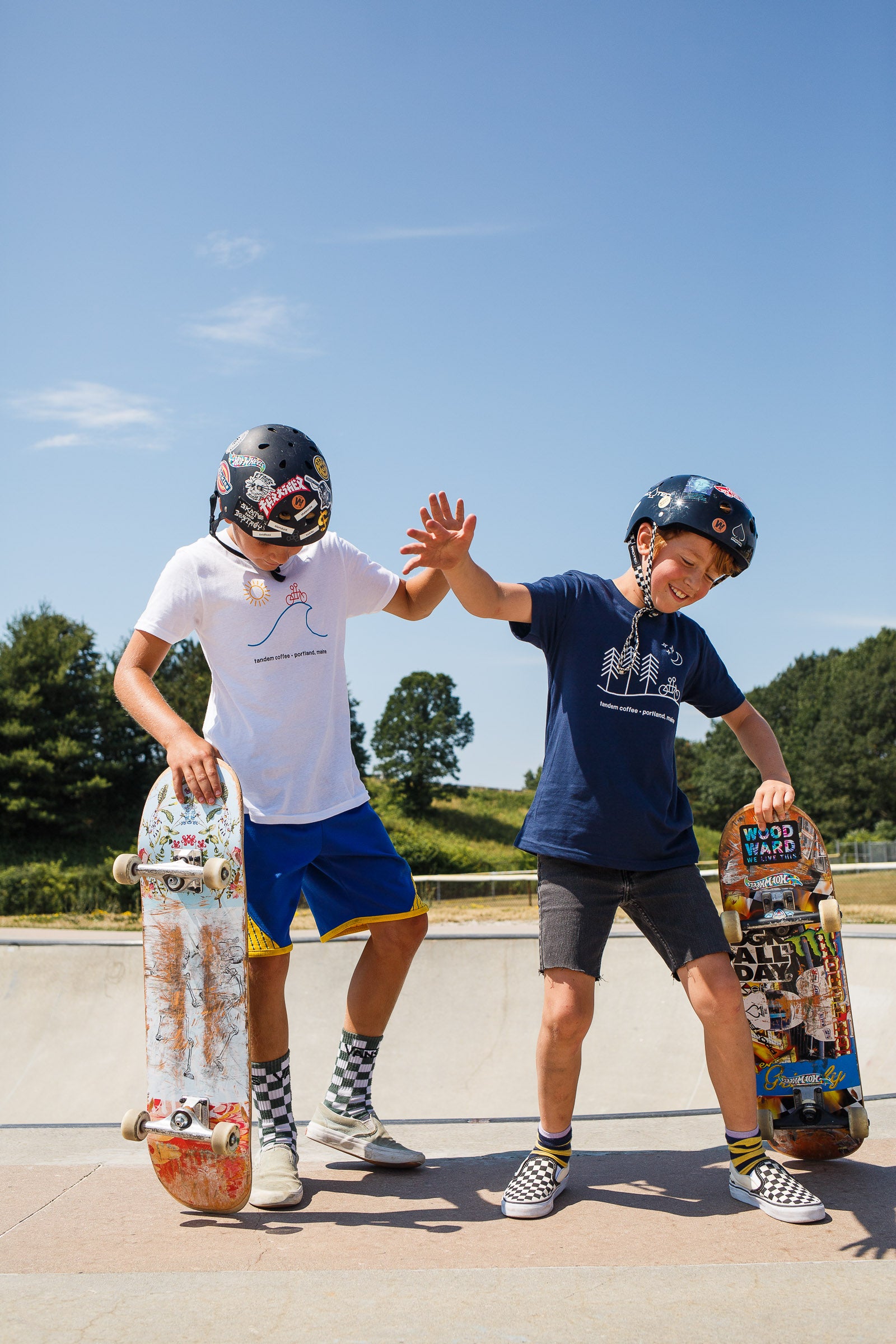 Two young boys in Tandem Kids Tandem Moon Tees and helmets stand at a skate park with skateboards, smiling and performing a high five under a clear blue sky.